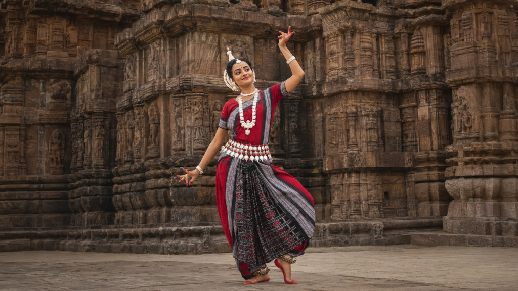 Odissi dancer performing in traditional costume in front of a carved temple wall