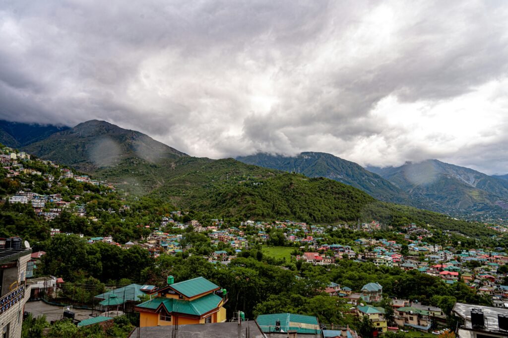 A Complete Dharamshala Travel Guide for First-Time Visitors 2 Panoramic view of Dharamshala with colourful hillside houses, lush green valleys and mountains beneath cloudy skies.