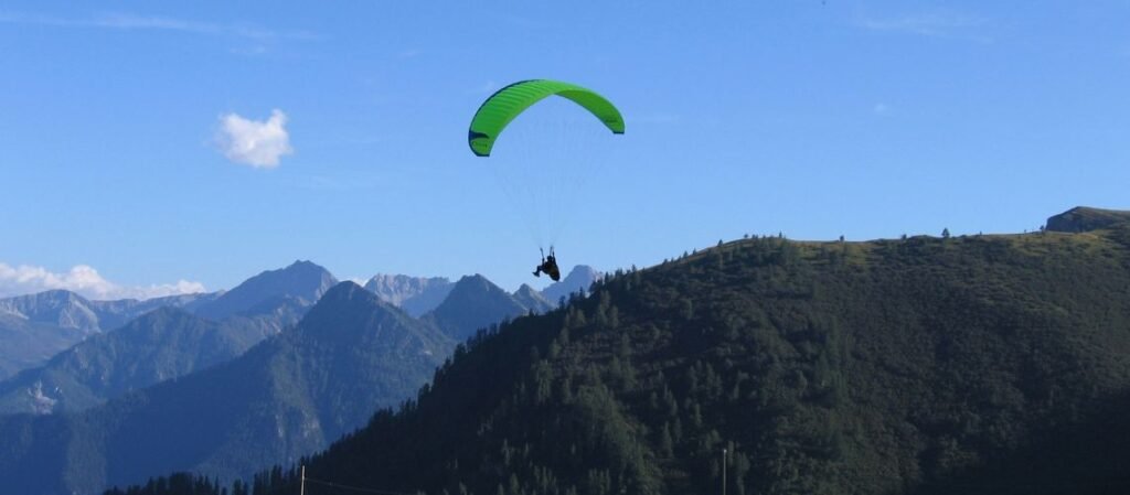 A scenic paragliding shot features a vibrant green canopy soaring over rugged mountain ridges under a clear blue sky.