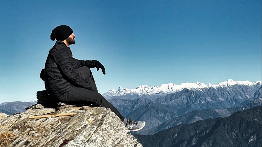 Traveller sitting on a rocky summit facing a snow-capped Himalayan range during a Shimla Manali tour package
